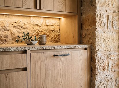 Detalle de cocina: frente de madera de roble, encimera de granito y muro de piedra caliza con iluminación rasante.