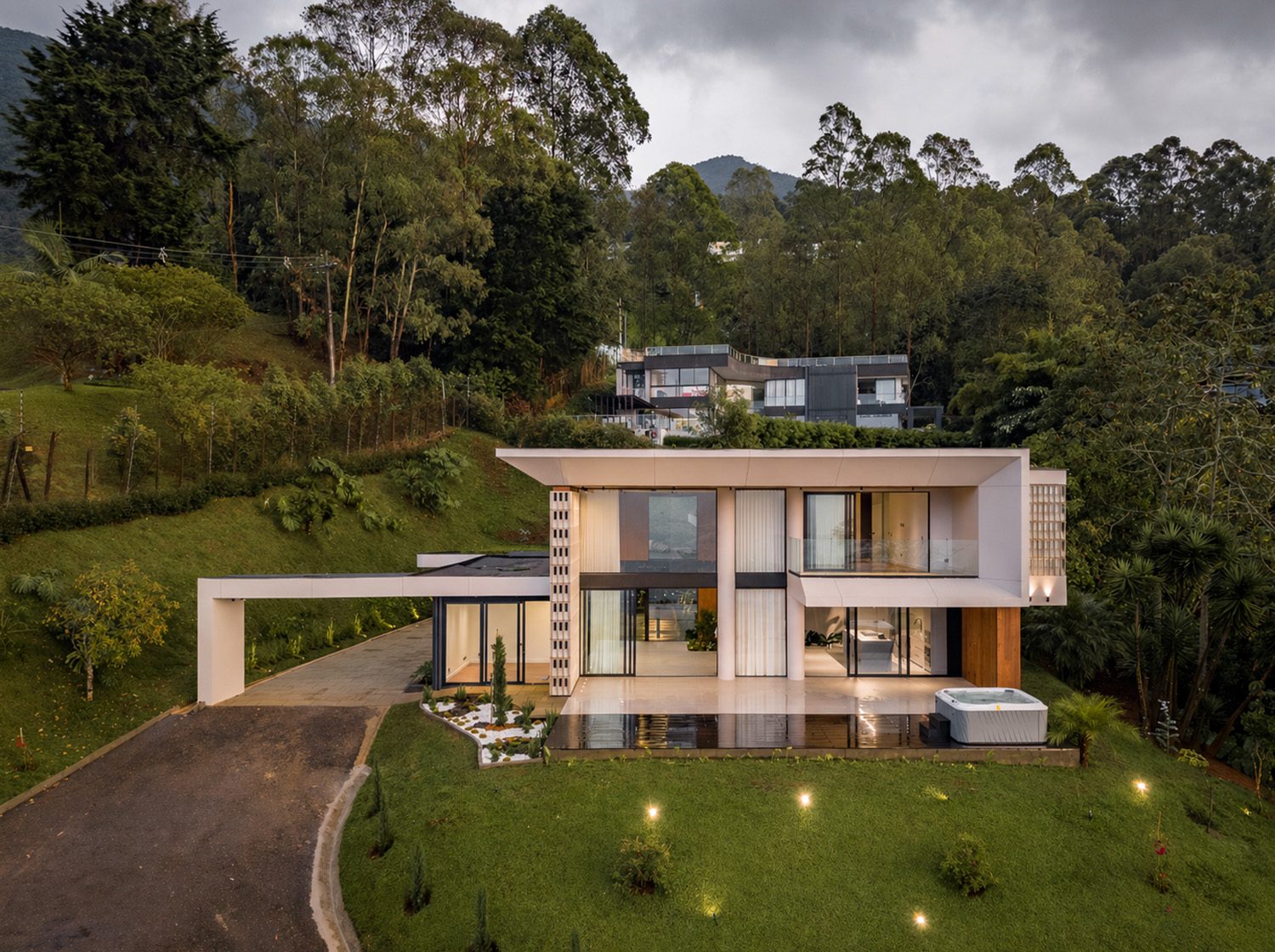 Casa Asturias al atardecer: volumen de dos plantas con fachada acristalada, jardín en pendiente y piscina, rodeado de bosque.