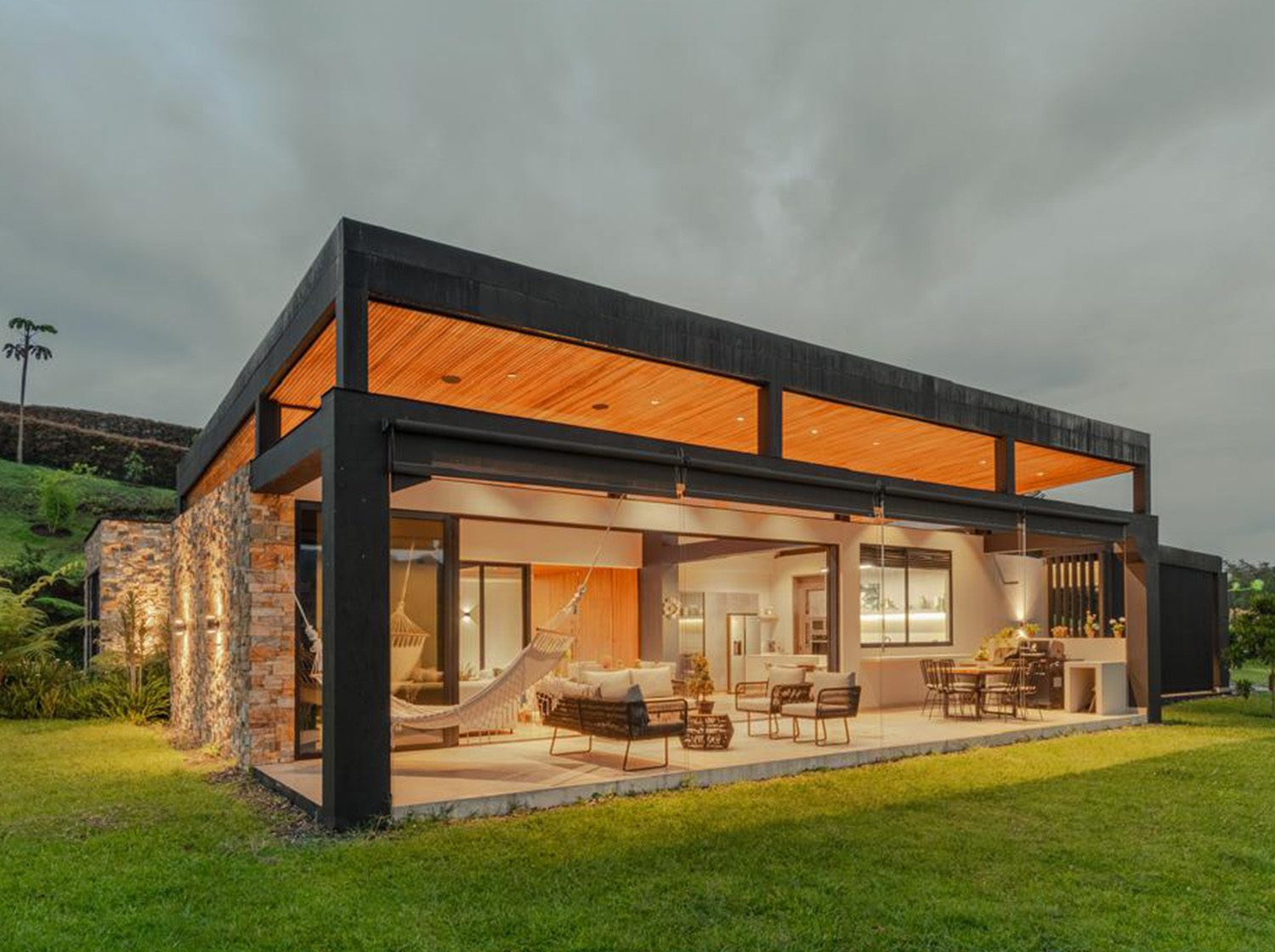 Casa LM: volumen de cubierta plana con estructura oscura y techo de madera, sobre un prado abierto.